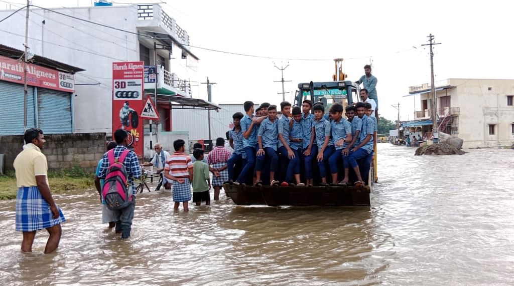 வெள்ளநீர் சாலையில் பெருக்கெடுத்து ஓடுவதால் பொக்லைன் இயந்திரத்தின் மீது அமர்ந்து சவாரி செய்யும் பள்ளி மாணவர்கள்.